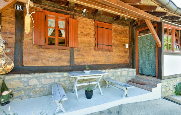 A rustic wooden building with a stone foundation, numbered 91. It features a wooden bench, a small table with a potted plant, and a set of steps leading to a door with a heart-shaped decoration.