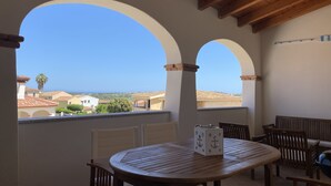 Um alpendre coberto com mesa e cadeiras de madeira, grandes janelas em arco com vista para o mar e edifícios ao redor.