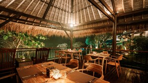 An outdoor dining area with wooden tables and chairs, thatched roof, and a view of the surrounding greenery. - One Degree Sunset Hill Leisure (Pecatu)