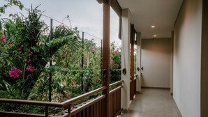 A corridor with a wooden railing, beige walls, and a view of lush greenery and pink flowers. - One Degree Sunset Hill Leisure (Pecatu)