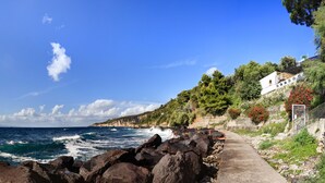 A coastal path with rocky terrain, a clear blue sky, and a building with a red railing on the right. - Appartamento Lobra (Massa Lubrense)