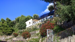 A hillside property with a white building featuring arched windows, surrounded by lush greenery and red flowering bushes. - Appartamento Lobra (Massa Lubrense)