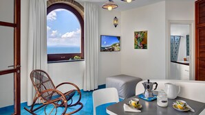 A room with a large window, a dining table with breakfast items, a rocking chair, and a view of the sea. - Appartamento Lobra (Massa Lubrense)