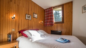 A bedroom with a wooden paneled wall, a bed with white bedding, a nightstand with books, a window with a red curtain, and a wall-mounted lamp. - Courchevel 1850: 2-room flat, south-facing balcony, parking (Courchevel 1850)