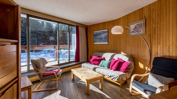 A cozy room with a wooden floor, a sofa with colorful cushions, a wooden coffee table, and a chair. There is a large window with a view of trees and a building outside. - Courchevel 1850: 2-room flat, south-facing balcony, parking (Courchevel 1850)