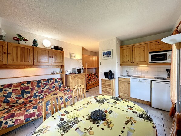 A compact kitchen with wooden cabinets, a microwave, and a dishwasher. There is a dining area with a patterned tablecloth and chairs.