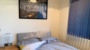 A bedroom with a bed, a nightstand, a lamp, a framed picture on the wall, and a window with blinds. - Cosy and Modern 2-bed House in Great Yarmouth (Great Yarmouth)