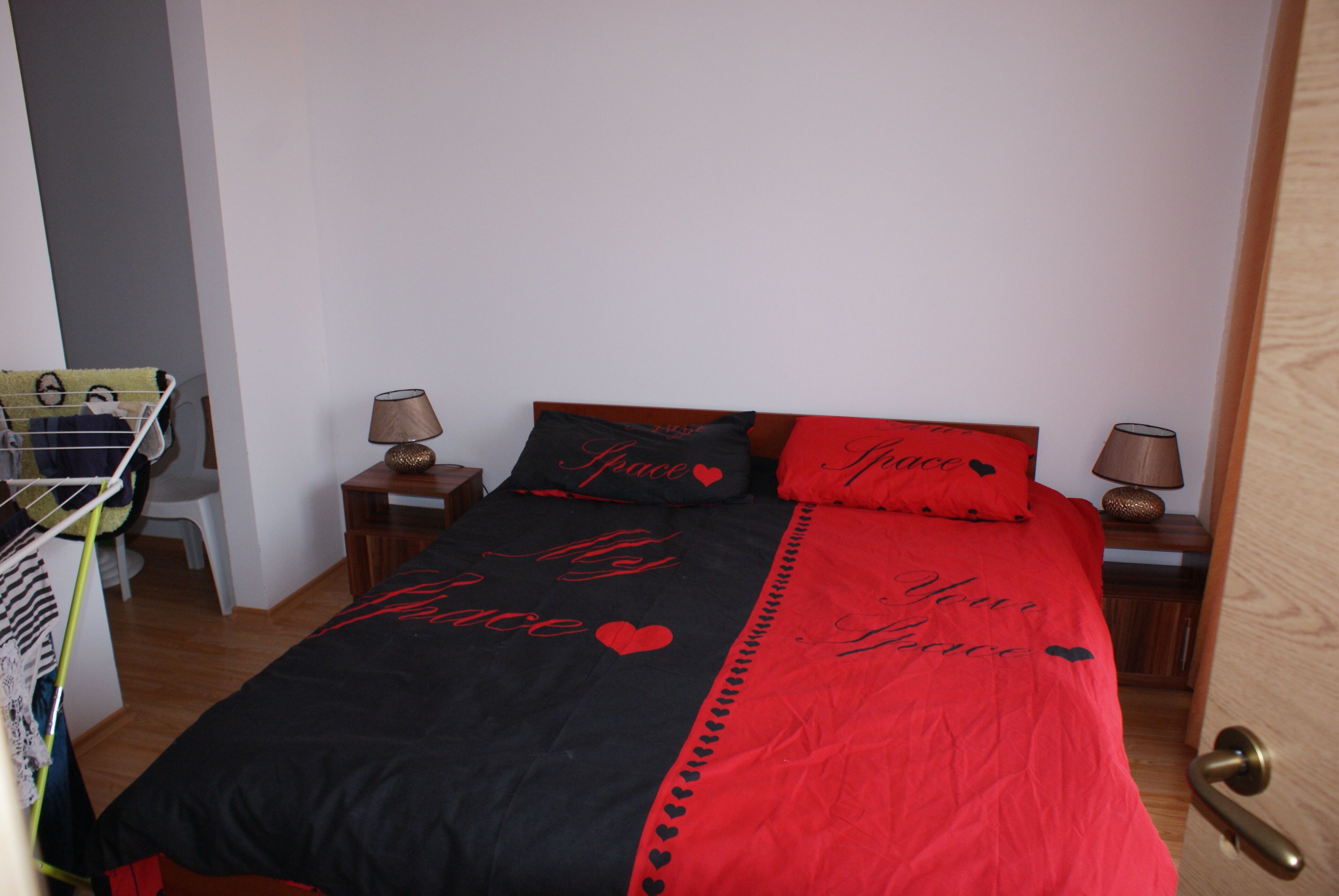 Un dormitorio con una cama cubierta con un edredón negro y rojo con la inscripción 'My Love', dos mesitas de noche con lámparas y una puerta de madera.