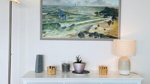 A framed painting of a seascape with a boat and sailboat on the water, a lamp, a candle, and a potted plant on a white dresser. - Elegant Apartment with Balcony, Garage and WiFi by the Sea (Les Sables-d'Olonne)