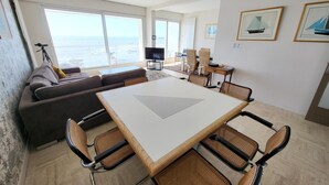 A modern living room with a sofa, dining table, and chairs, large windows with a view of the sea, and framed pictures on the wall. - Elegant Apartment with Balcony, Garage and WiFi by the Sea (Les Sables-d'Olonne)