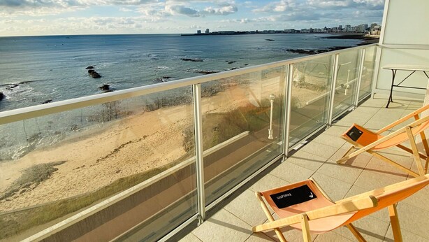 A balcony with a glass railing overlooking a beach. There are two wooden lounge chairs with cushions. - Elegant Apartment with Balcony, Garage and WiFi by the Sea (Les Sables-d'Olonne)