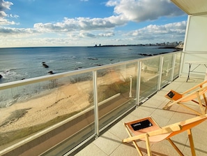 A balcony with a glass railing overlooking a beach. There are two wooden lounge chairs with cushions.