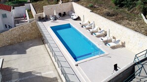 A rectangular swimming pool with lounge chairs and umbrellas, surrounded by stone walls and a white building with a red roof. - Villa Samba (Zupa dubrovacka)