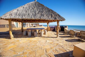A thatched-roof outdoor seating area with stone flooring, overlooking the sea.