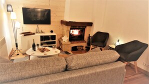 A cozy living room with a fireplace, a flat-screen TV, a sofa, and a dining table with wine glasses and a bottle. - 4* family apartment, terrace, garden and fireplace (Briançon)