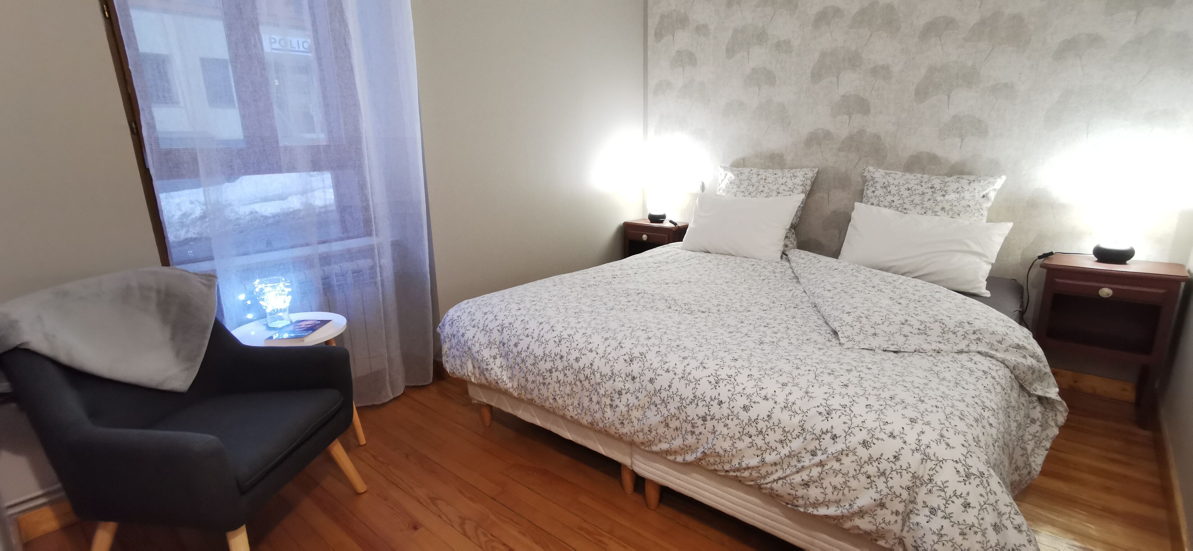 A bedroom with a bed, two bedside tables, a chair, a small table, and a window with curtains.