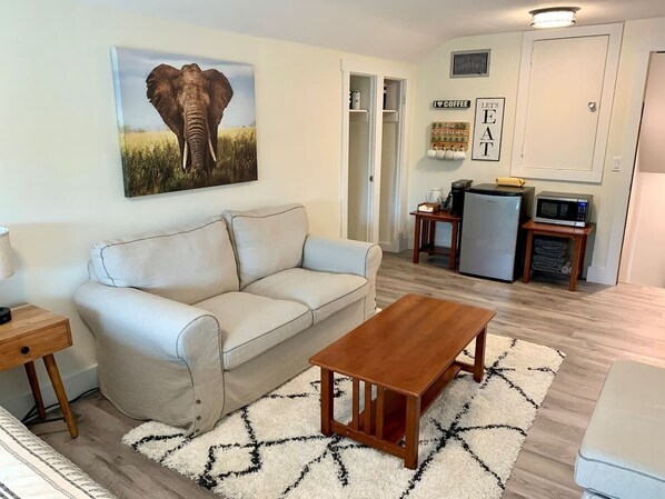 A living room with a sofa, coffee table, and a painting of an elephant. There is a kitchen area with a refrigerator, microwave, and a coffee station.