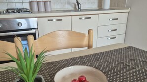 Una cocina con estufa, horno y armarios. Hay una mesa con mantel estampado y un tazón con objetos rojos. Una planta en maceta está sobre la mesa.