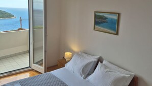 Un dormitorio con cama, mesita de noche, lámpara, un cuadro y un ventanal corredero que da a un balcón con vistas al mar y una isla distante.