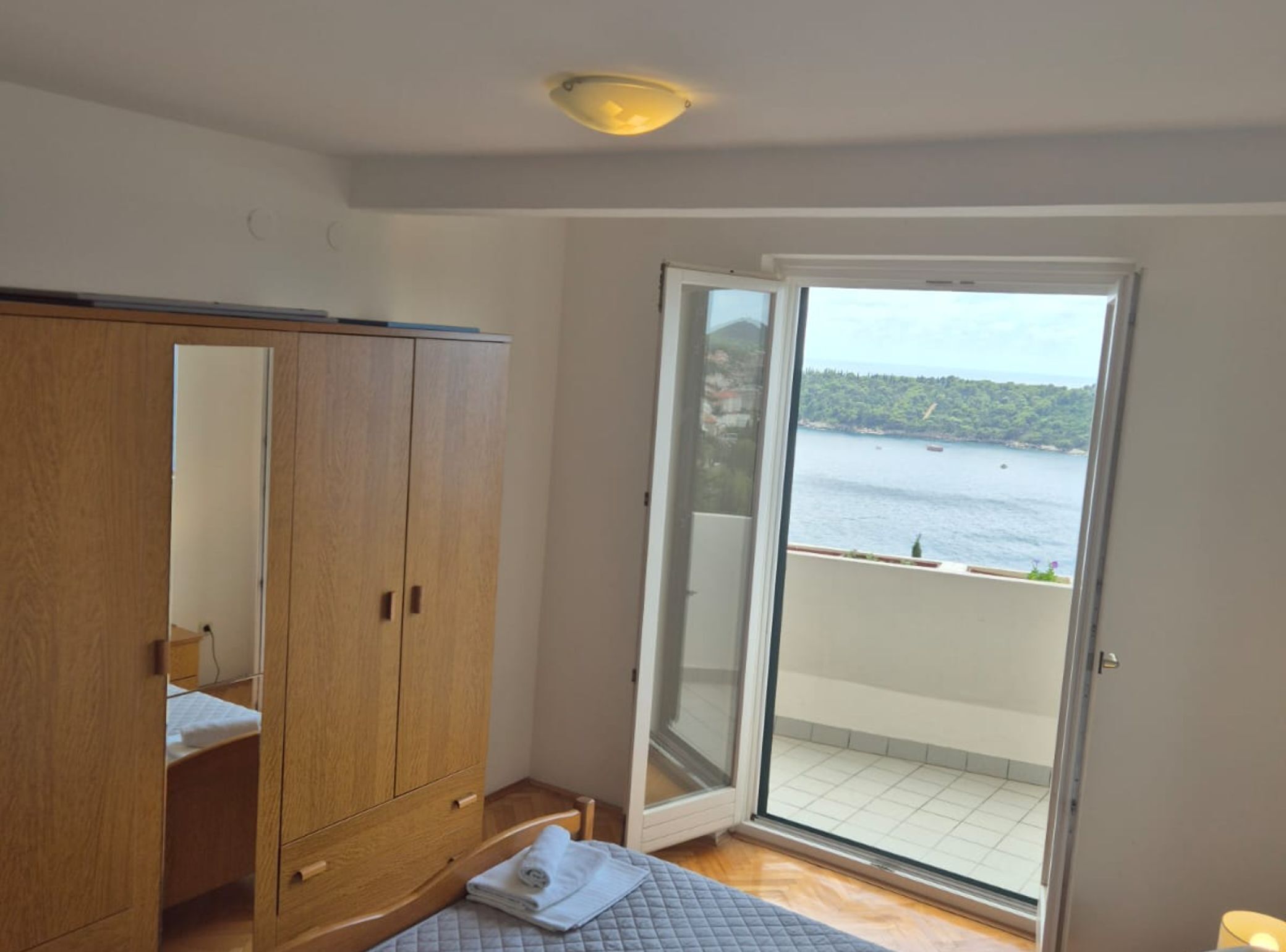 Una camera da letto con un'armadio in legno, un letto con copriletto e una porta scorrevole in vetro che dà su un balcone con vista sull'acqua.