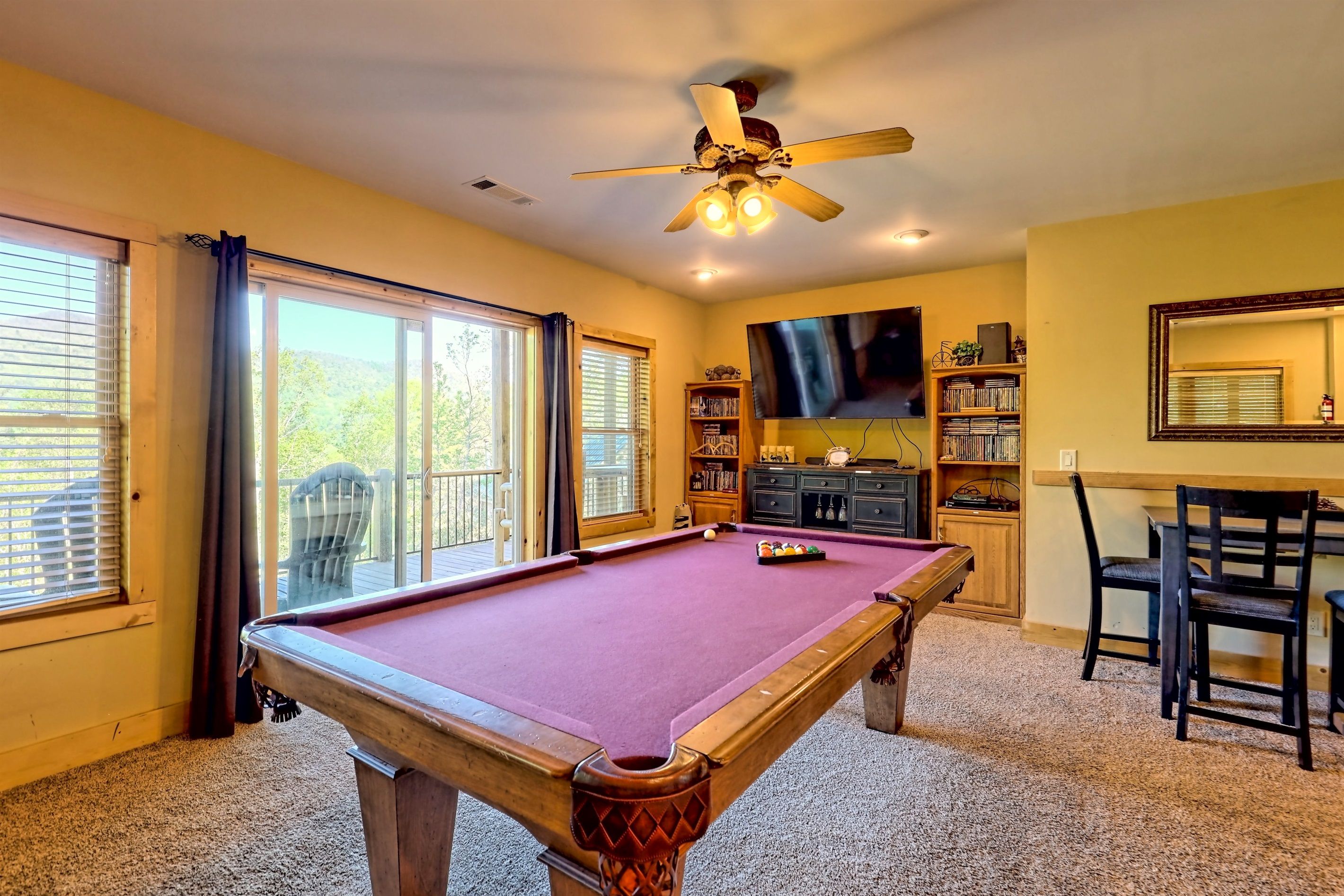 A pool table in a room with a ceiling fan, large windows, a bookshelf, and a TV.