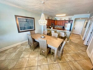 A dining area with a wooden table and wicker chairs, a chandelier, and a kitchen in the background.