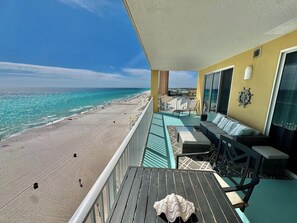 A balcony with a view of the beach and ocean, a sofa, a table, and a ship wheel decoration.