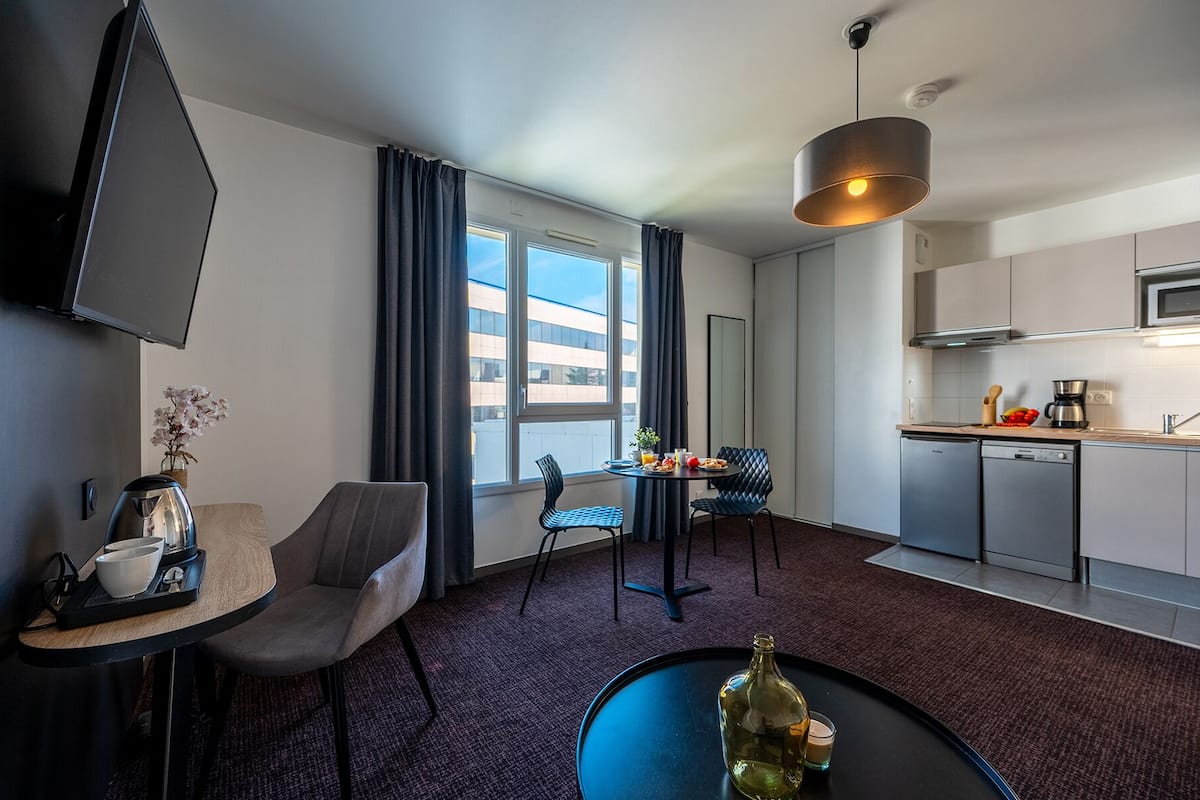 Une chambre d’hôtel moderne dotée d’un coin repas, d’une cuisine et d’une grande fenêtre donnant sur la ville.
