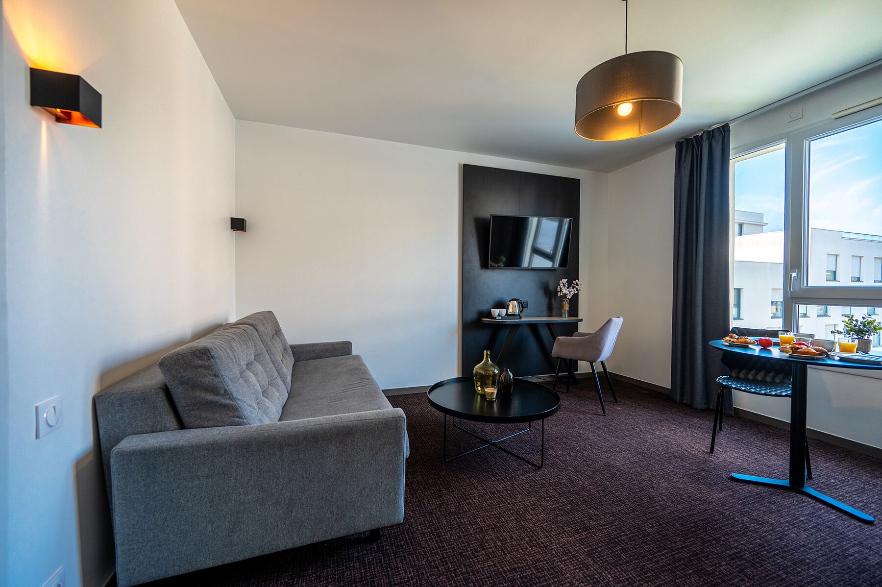 A modern hotel room with a grey sofa, a small round table, a TV mounted on the wall, and a window with a view of the city.