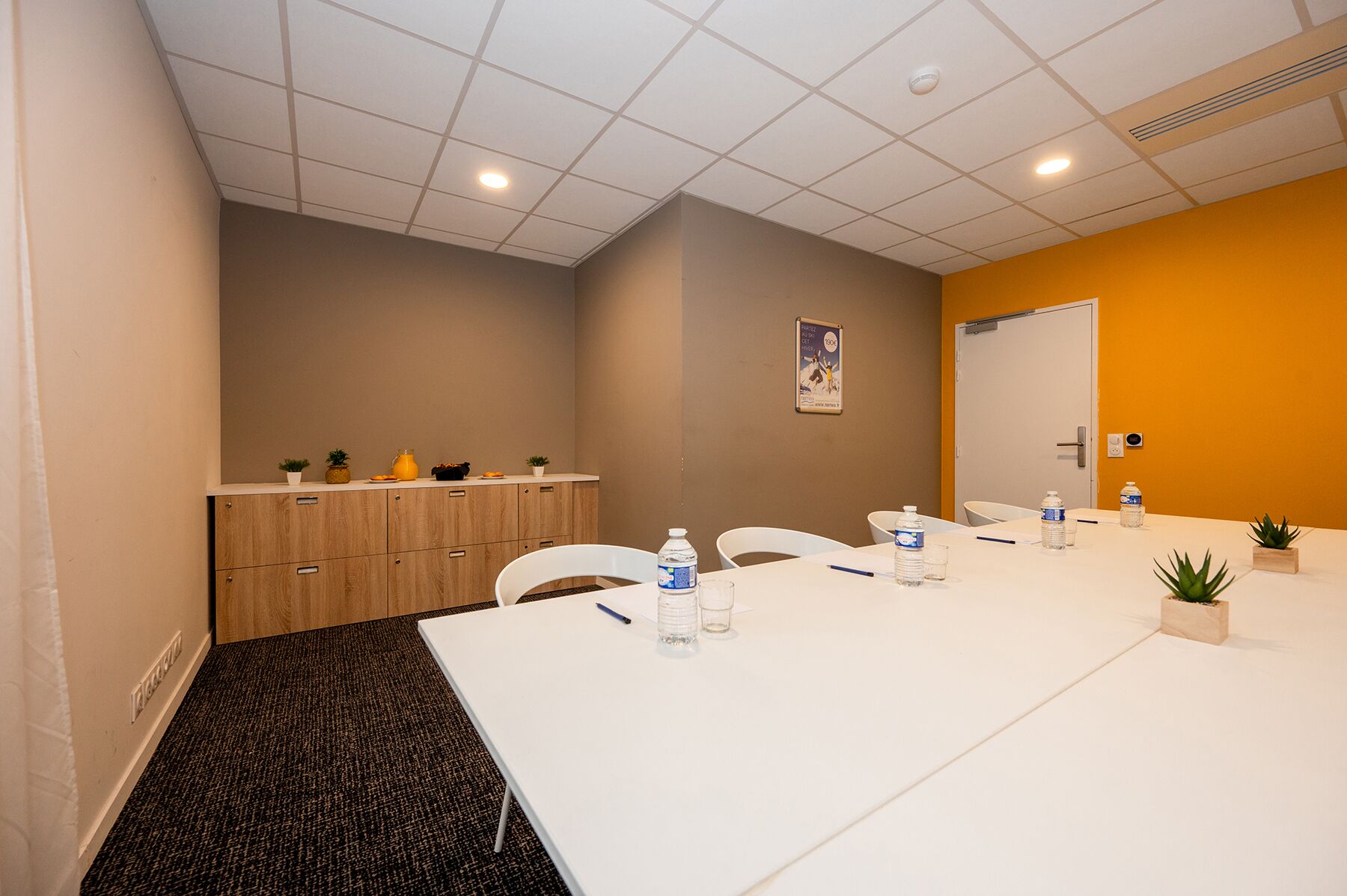 Een vergaderruimte met een witte tafel, stoelen, een waterdispenser en een kleine plant.