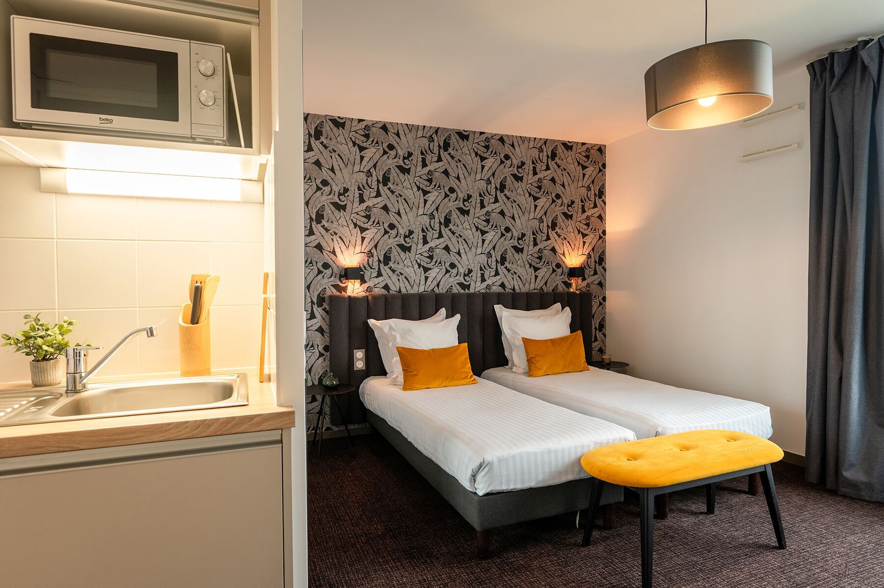 Une chambre d’hôtel compacte comprenant un coin cuisine, un lit avec deux oreillers, une table de chevet et un luminaire suspendu.