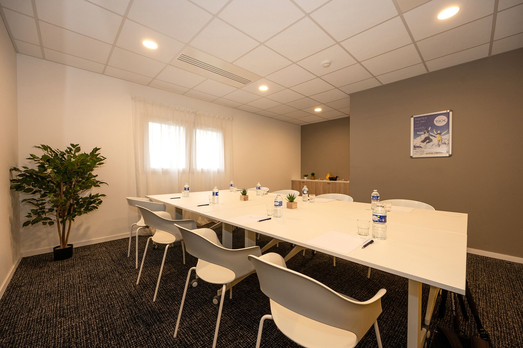 Une salle de conférence avec une longue table, des chaises blanches, des bouteilles d’eau et une plante en pot.