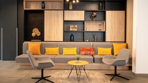 A modern living room with a grey sofa, two armchairs, a round coffee table, and a wooden shelving unit with decorative items. -  Nemea Appart Hotel Europe Velizy Villacoublay (Velizy-Villacoublay)