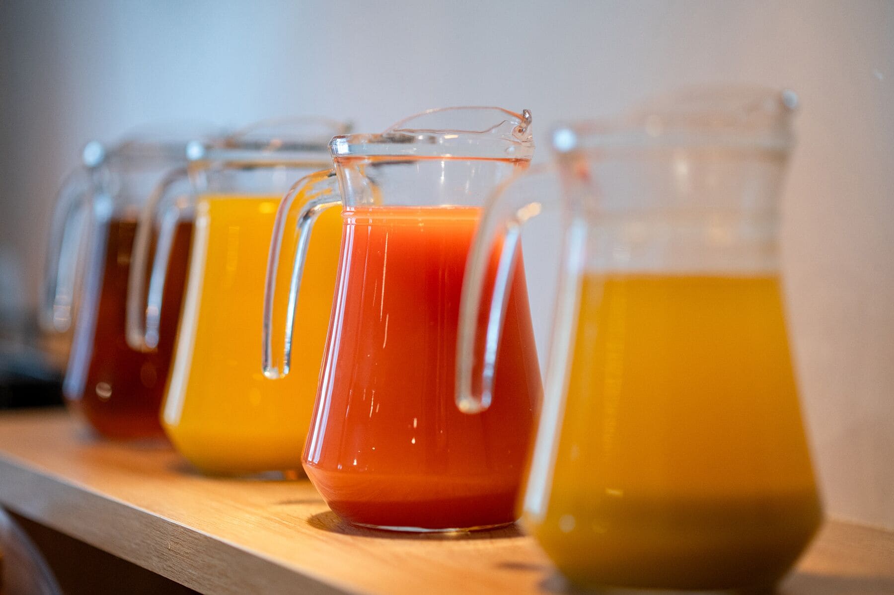 A row of glass pitchers with handles, containing different colored liquids, likely juices or smoothies, arranged on a wooden surface.