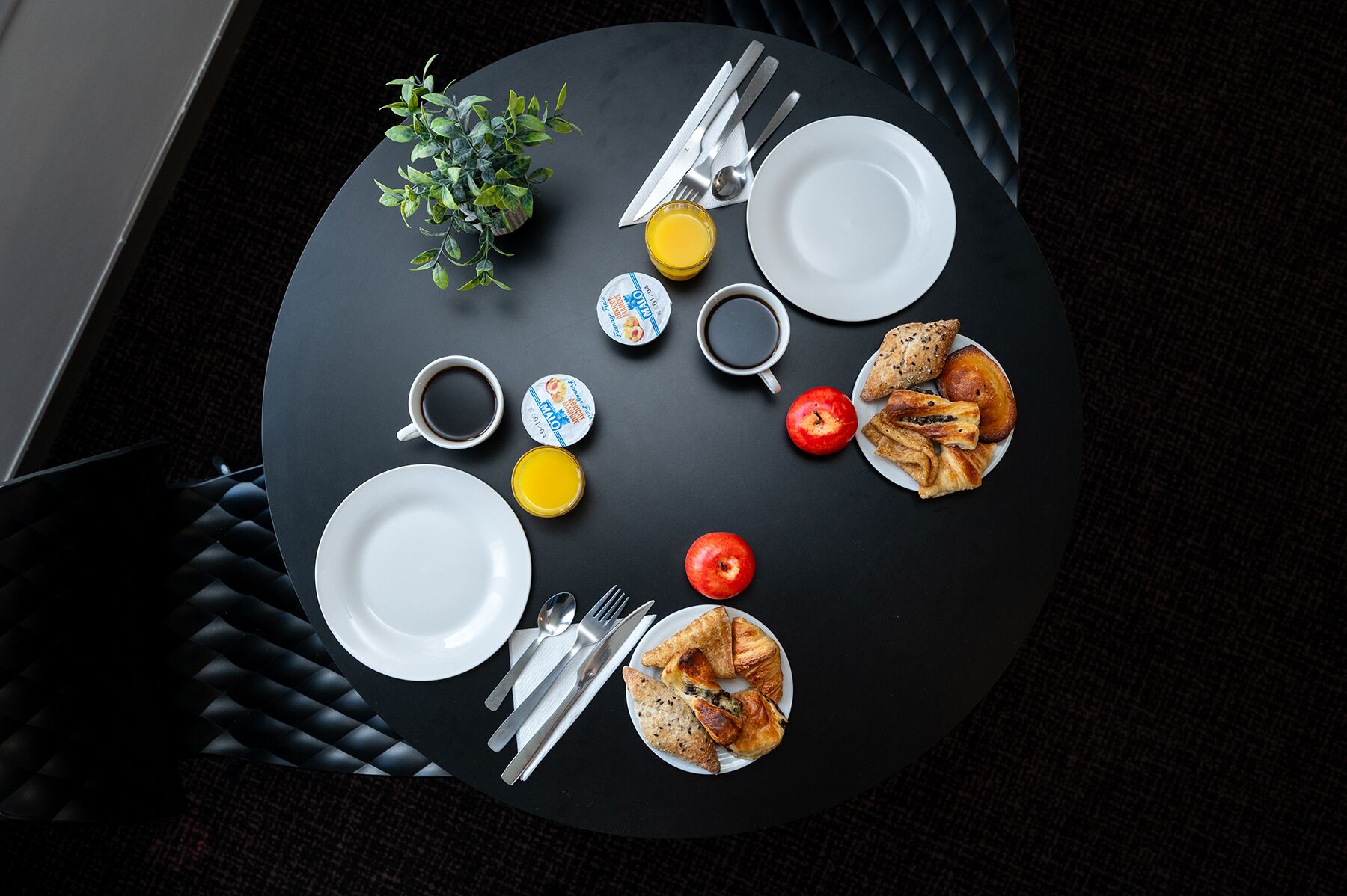 A round dining table with plates, cutlery, a glass of juice, a cup of coffee, a small plant, and a plate of pastries.