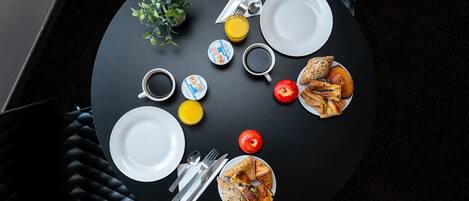 A round dining table with plates, cutlery, a glass of juice, a cup of coffee, a small plant, and a plate of pastries.