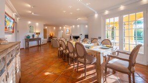 Une salle à manger spacieuse, avec une grande table dressée pour six personnes, entourée de six chaises. La pièce est dotée d’un sol carrelé, de murs blancs et d’une grande fenêtre donnant sur un paysage verdoyant.