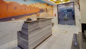 A hotel reception area with a marble counter, a mural on the wall, and a glass elevator door. - Jasoda Heritage Hotel Near Hawa Mahal (Jaipur)