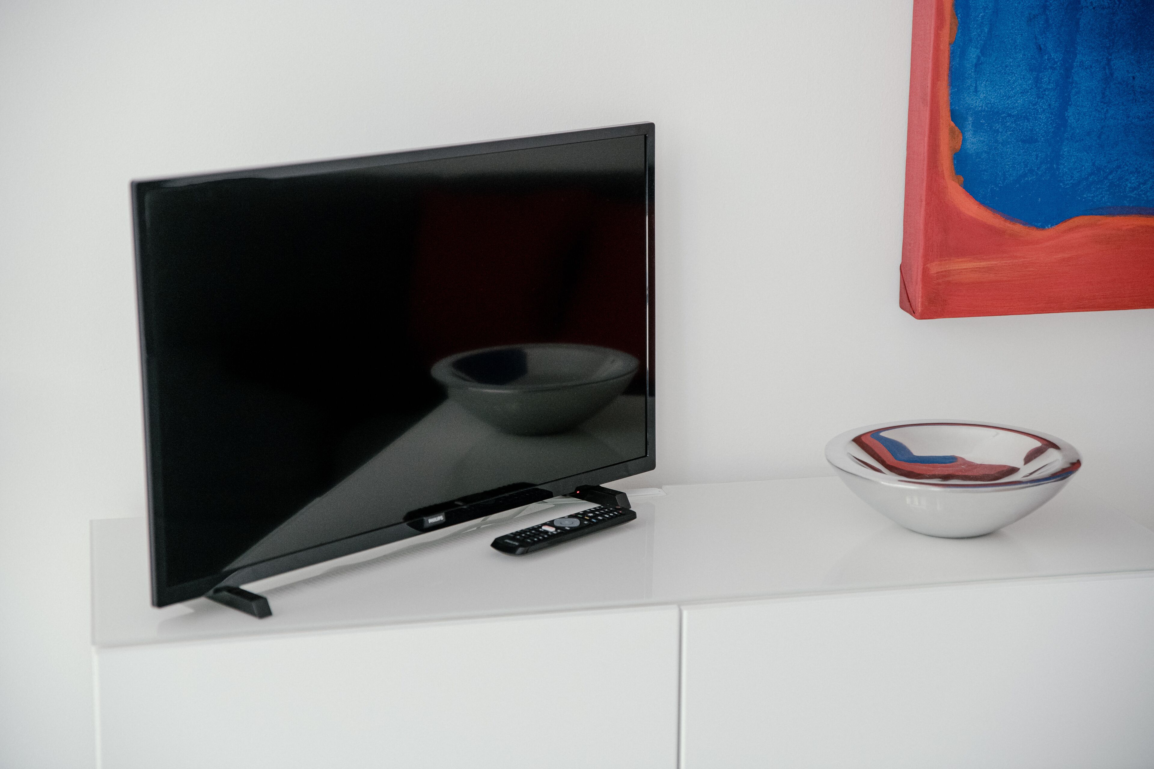 Un téléviseur à écran plat posé sur un meuble blanc, avec une télécommande et un bol.
