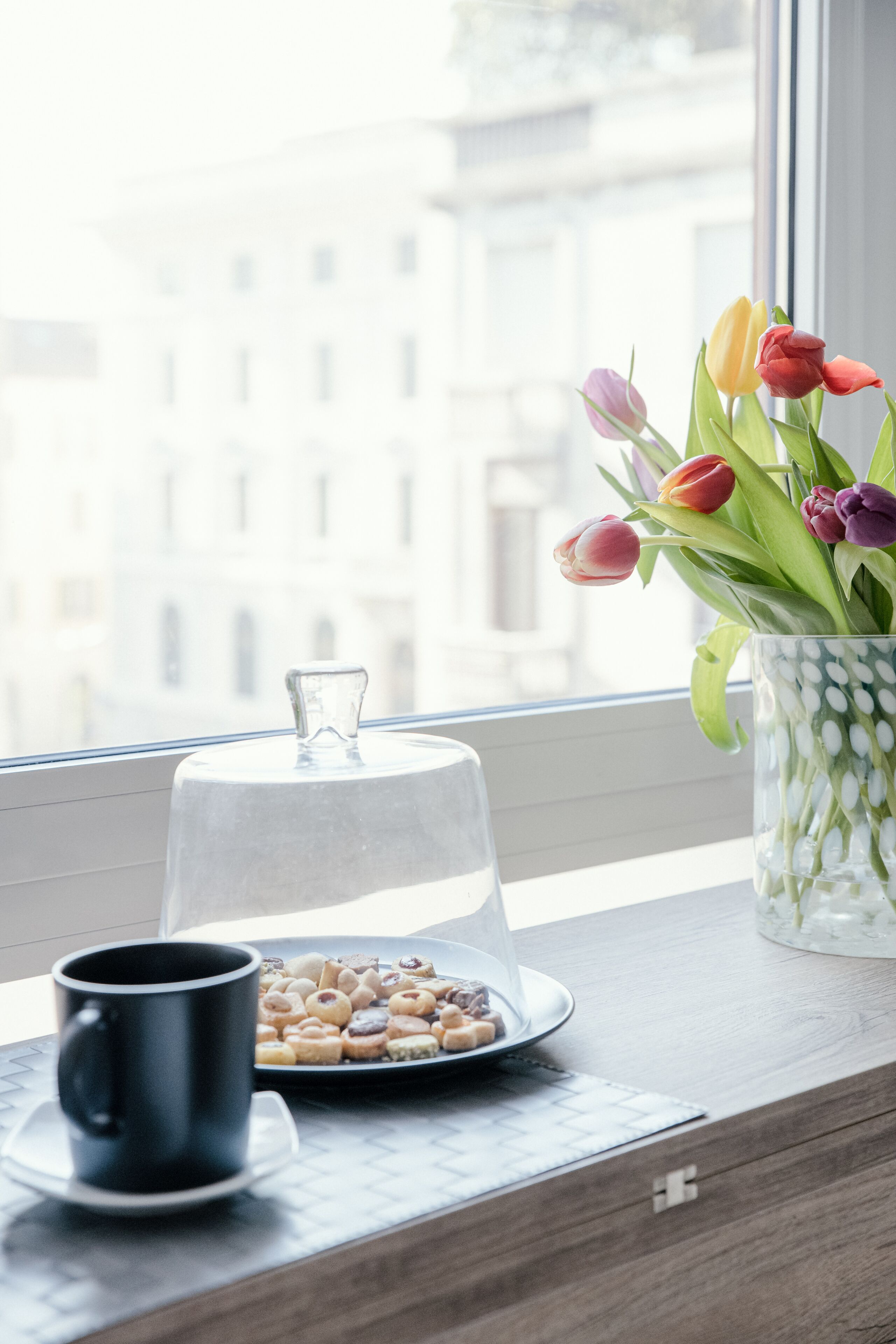Blick aus dem Fenster mit einer Tulpenvase auf einem Fensterbrett, einer Glasglocke und einer Auswahl an Gebäck.