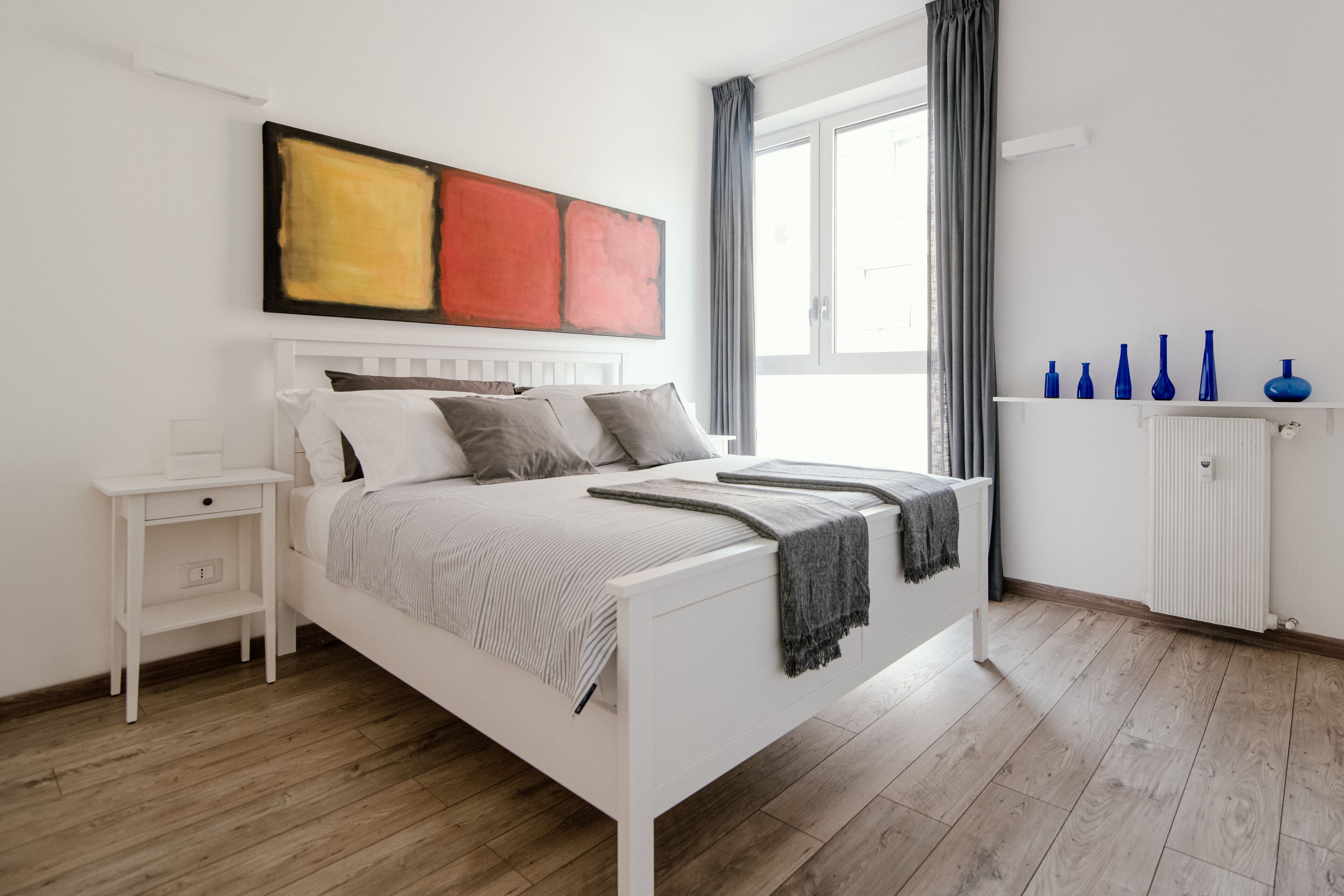 Un dormitori modern amb un llit blanc, roba de llit gris, un quadre abstracte gran, una finestra amb cortines i un radiador blanc amb gerros blaus.