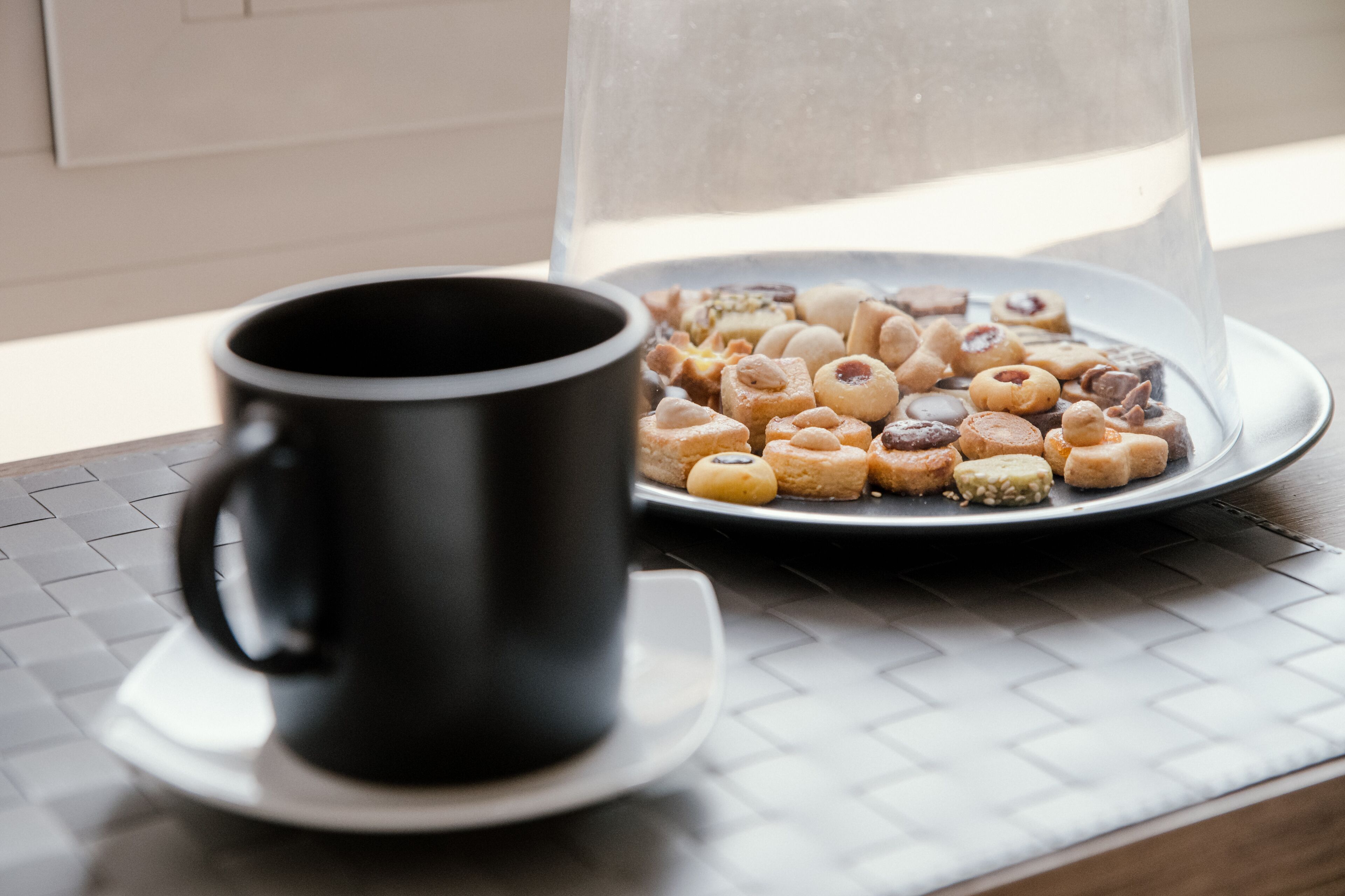 A black coffee cup on a saucer next to a plate of assorted pastries under a glass dome.
