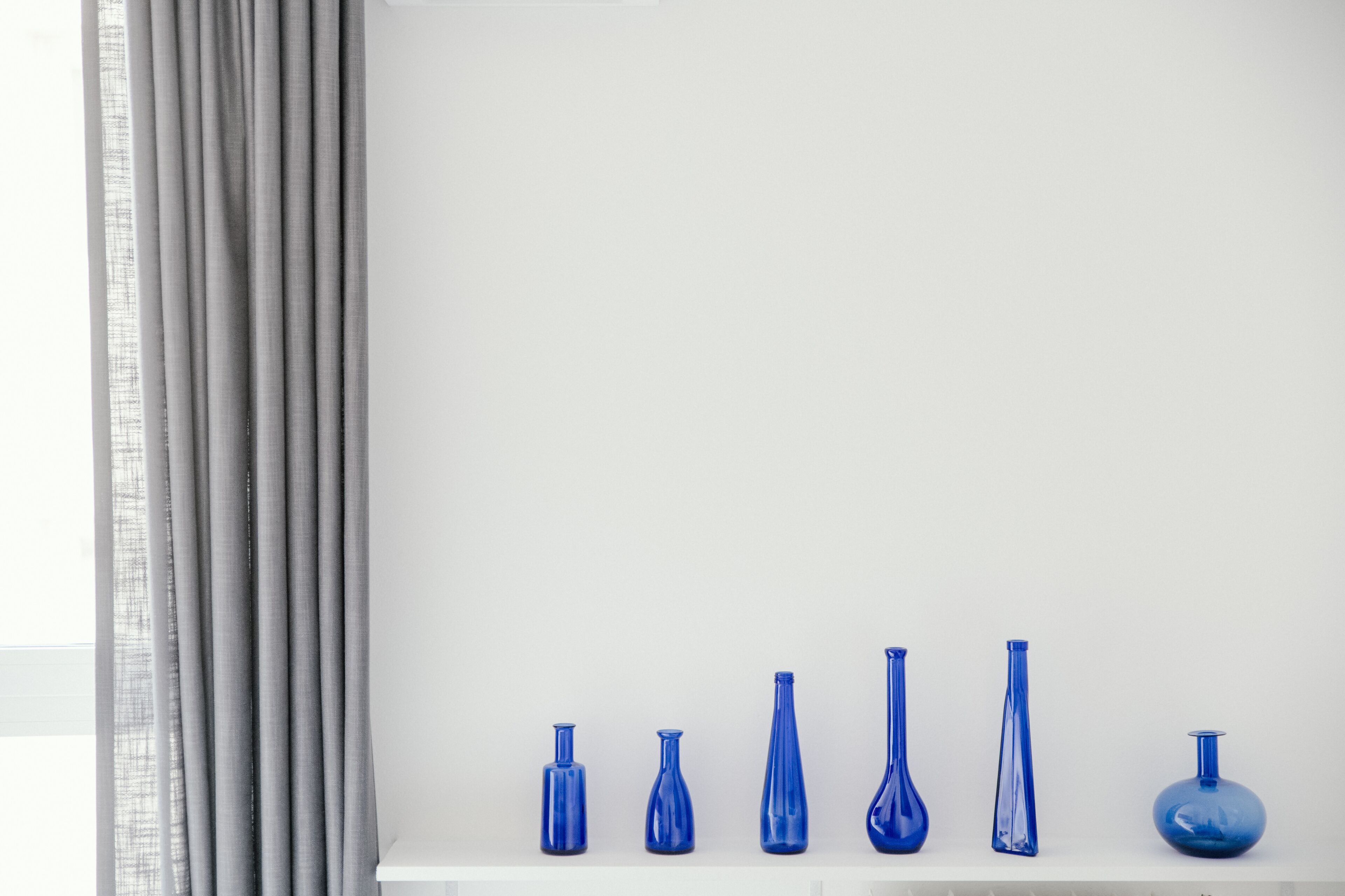 Une étagère blanche sur laquelle sont disposés six vases en verre bleu de formes différentes, le tout adossé à un mur blanc.