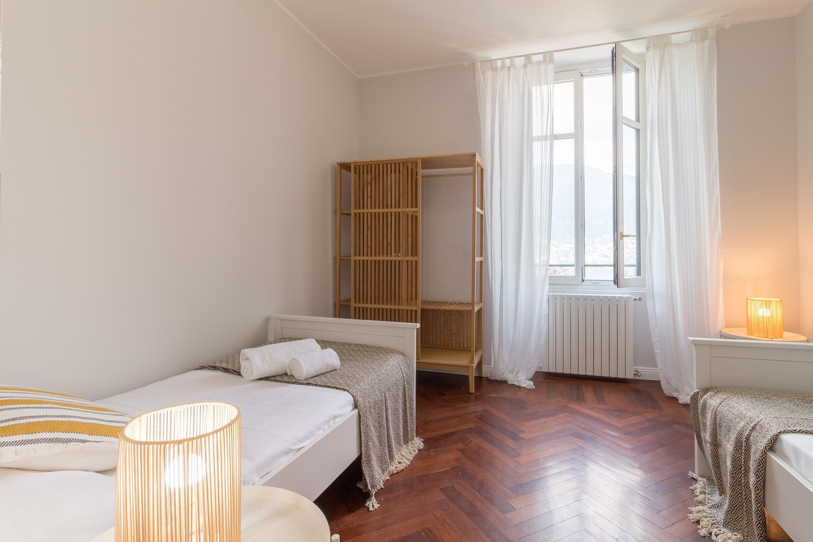 A bedroom with two single beds, a wooden wardrobe, a radiator, and a window with sheer curtains.