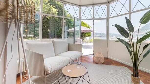 A modern, well-lit room with a white sofa, a small round table, and a large potted plant. - Como Lake Vista by My Home in Como (Blevio)