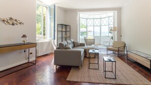 A modern living room with a sofa, a coffee table, and a side table. There is a large window with a view of greenery outside. - Como Lake Vista by My Home in Como (Blevio)