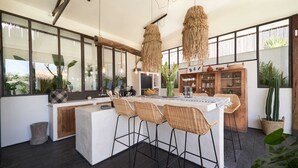 A modern kitchen with a central island, bar stools, hanging light fixtures, and large windows overlooking a garden. - Fabulous 3BR Boho-chic Style, Umalas With Staff (Kerobokan)