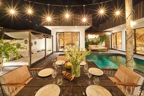 An outdoor dining area with a pool, wooden furniture, and string lights. - Fabulous 3BR Boho-chic Style, Umalas With Staff (Kerobokan)