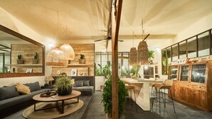 A modern living room with a sofa, coffee table, and hanging lights. There is a kitchen area with bar stools and a dining table. - Fabulous 3BR Boho-chic Style, Umalas With Staff (Kerobokan)