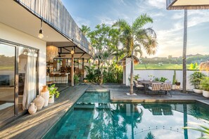 A modern outdoor pool area with a dining table and chairs, surrounded by potted plants and palm trees. - Fabulous 3BR Boho-chic Style, Umalas With Staff (Kerobokan)